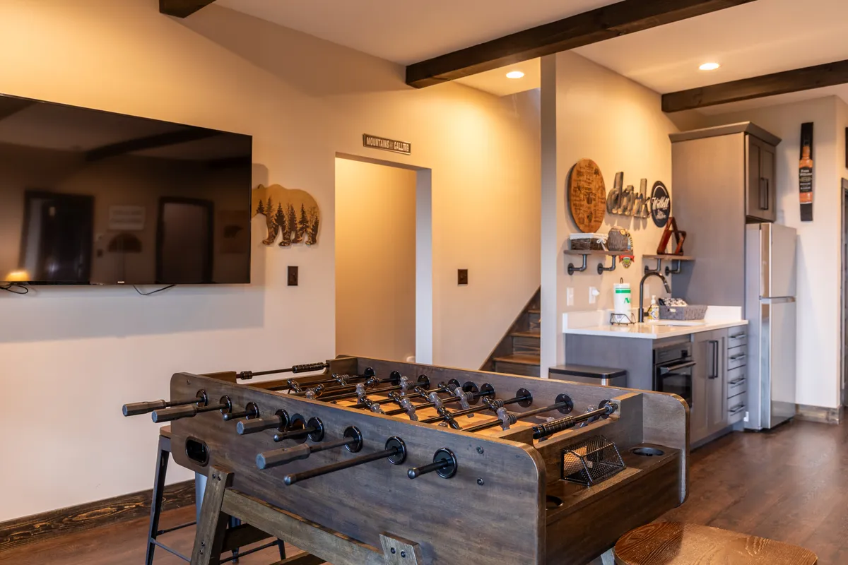 A lively area with a foosball table in the foreground, a flat-screen TV on the wall, and a kitchenette nearby, creating a playful and interactive setting ideal for friendly competition and group entertainment.