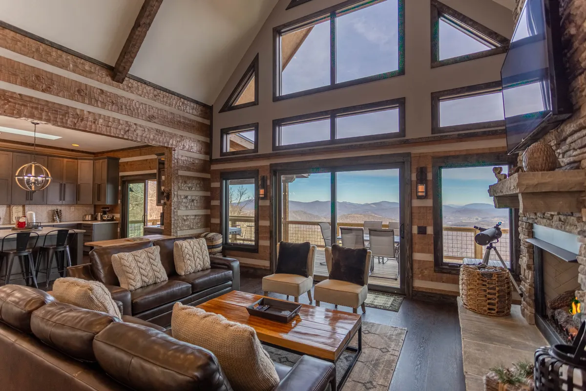 A spacious great room featuring comfortable leather sofas arranged around a stunning stacked stone fireplace. Floor-to-ceiling windows flood the space with natural light and offer breathtaking, unobstructed mountain views. The wooden coffee table and rustic accents create a warm, welcoming setting ideal for family relaxation and entertaining guests.