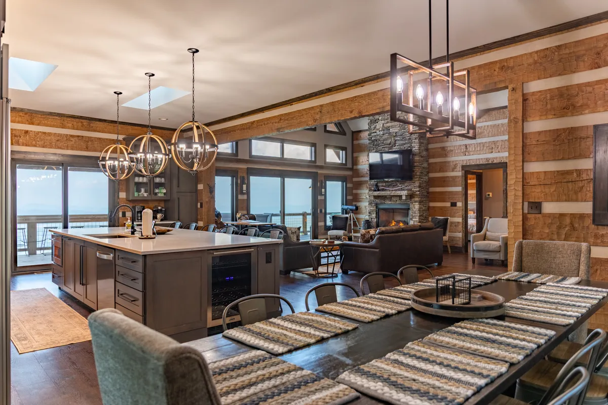 An open-concept living area combining the chef’s kitchen, dining table for ten, and a cozy stone fireplace under soaring wooden-beamed ceilings. Large windows provide panoramic mountain views, while the kitchen island and dining setting invite guests to cook, dine, and socialize together.