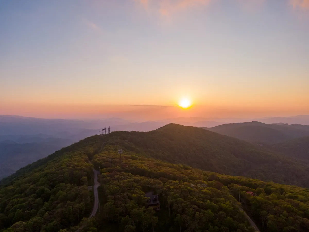 A breathtaking sunset casts golden and orange hues over distant mountain peaks, dense forests, and a winding road that cuts through the landscape. The scene is tranquil yet invigorating, hinting at the outdoor possibilities that await, with visible opportunities for hiking and nature photography.