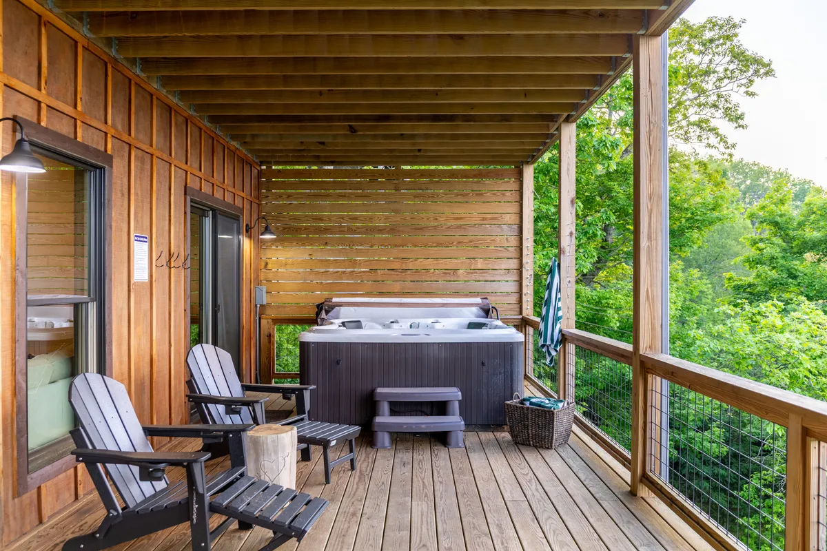A large wooden deck featuring a 7-person hot tub surrounded by Adirondack chairs, set against a backdrop of forest and mountains. The deck is positioned to take advantage of sunset views, combining luxury and natural beauty.