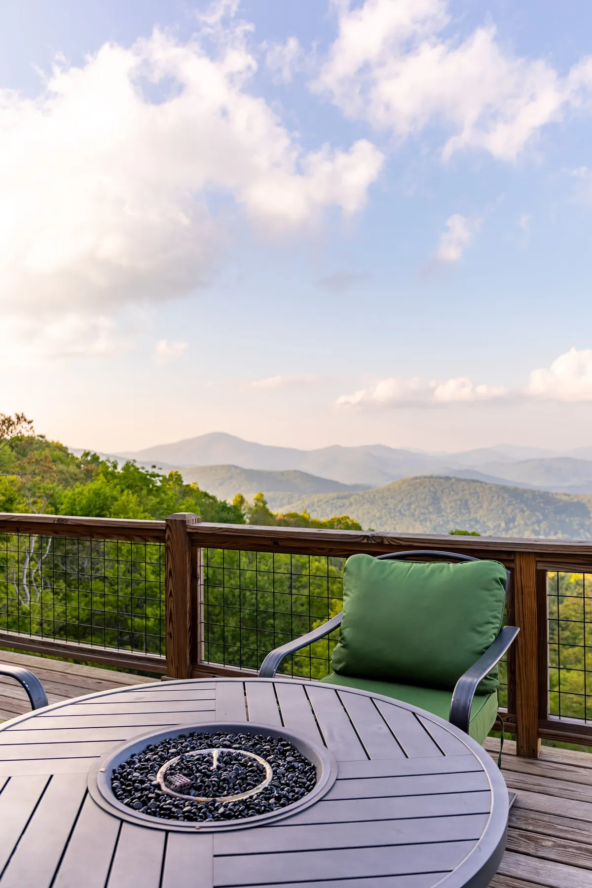 An inviting exterior scene featuring a fire pit surrounded by green chairs on a patio, set against a sweeping backdrop of mountains. The area encourages socializing and enjoying nature, ideal for stargazing, morning coffee, or evening conversations with loved ones.