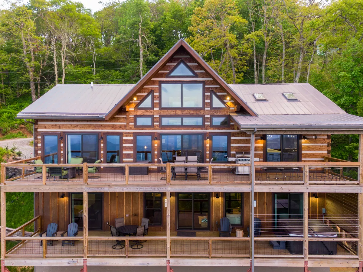 The image features the stunning exterior of a newly built luxury log cabin nestled among tall trees, with expansive glass windows and a spacious deck overlooking layered mountain vistas. The wooden structure exudes warmth and elegance, while the deck is set up for outdoor gatherings, relaxation, and grilling, all framed by the natural beauty of Boone, North Carolina.