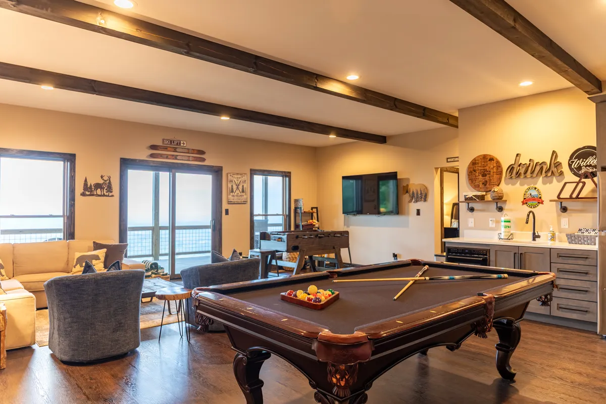 A spacious game room featuring a pool table at the center, a comfortable sofa along one wall, a large TV mounted nearby, and a well-equipped bar area with a refrigerator and microwave. The space is designed for both play and relaxation, with ample room for gatherings.