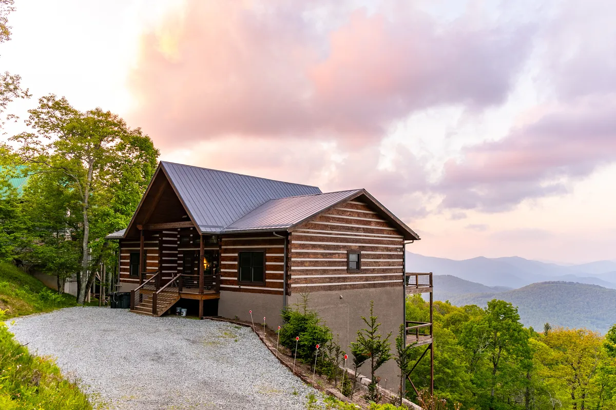 The log cabin's rustic architecture is set against a dramatic mountain backdrop and a glowing sunset sky. The exterior view captures the remote, tranquil setting, inviting couples to explore the area or simply soak in the scenery from the home itself.