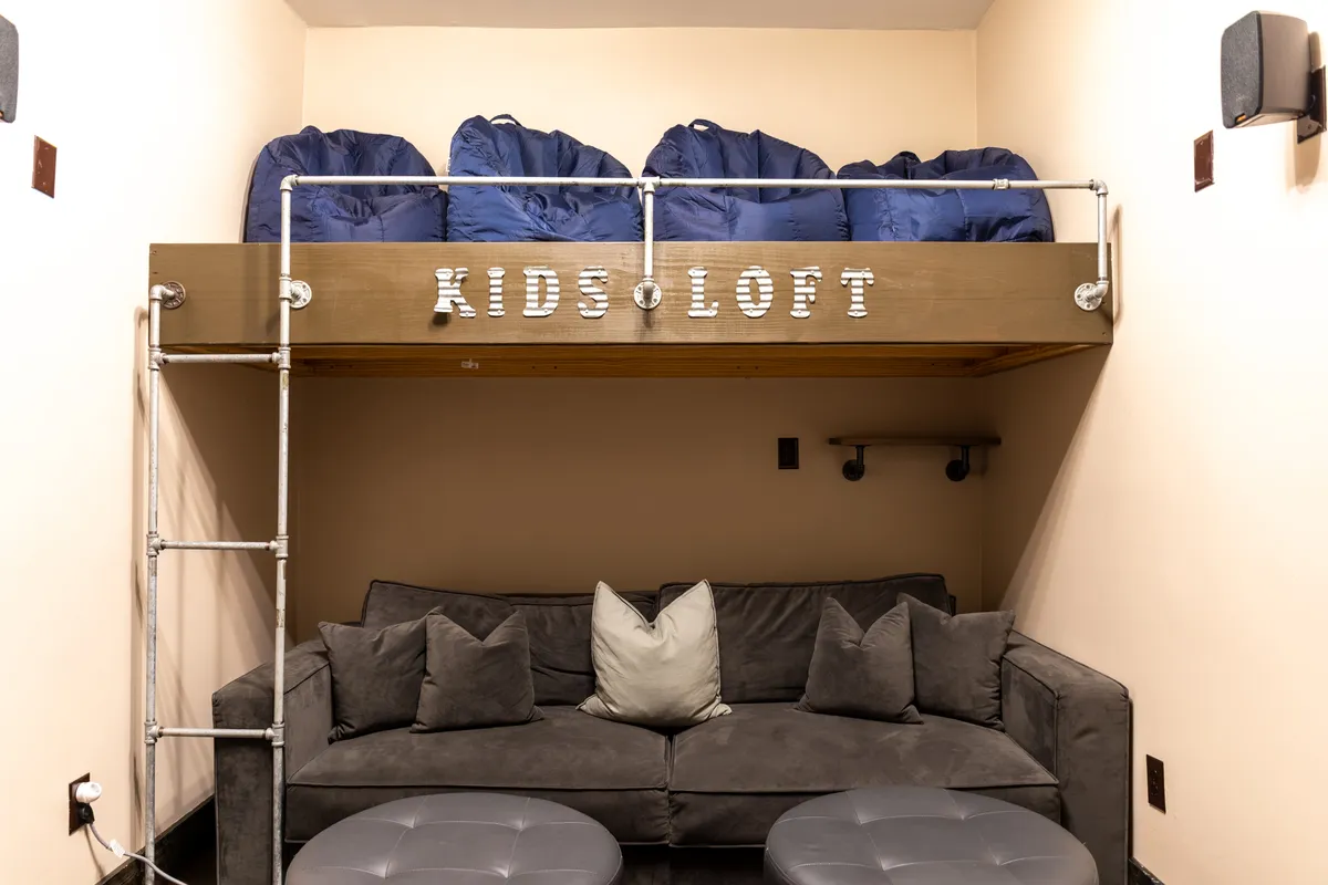 A playful, lofted interior space designed for kids, featuring bean bags, a comfortable couch, and decorative letters spelling 'KIDS LOFT.' The space is cozy, inviting, and clearly dedicated to younger guests for reading, playing games, and imaginative activities.