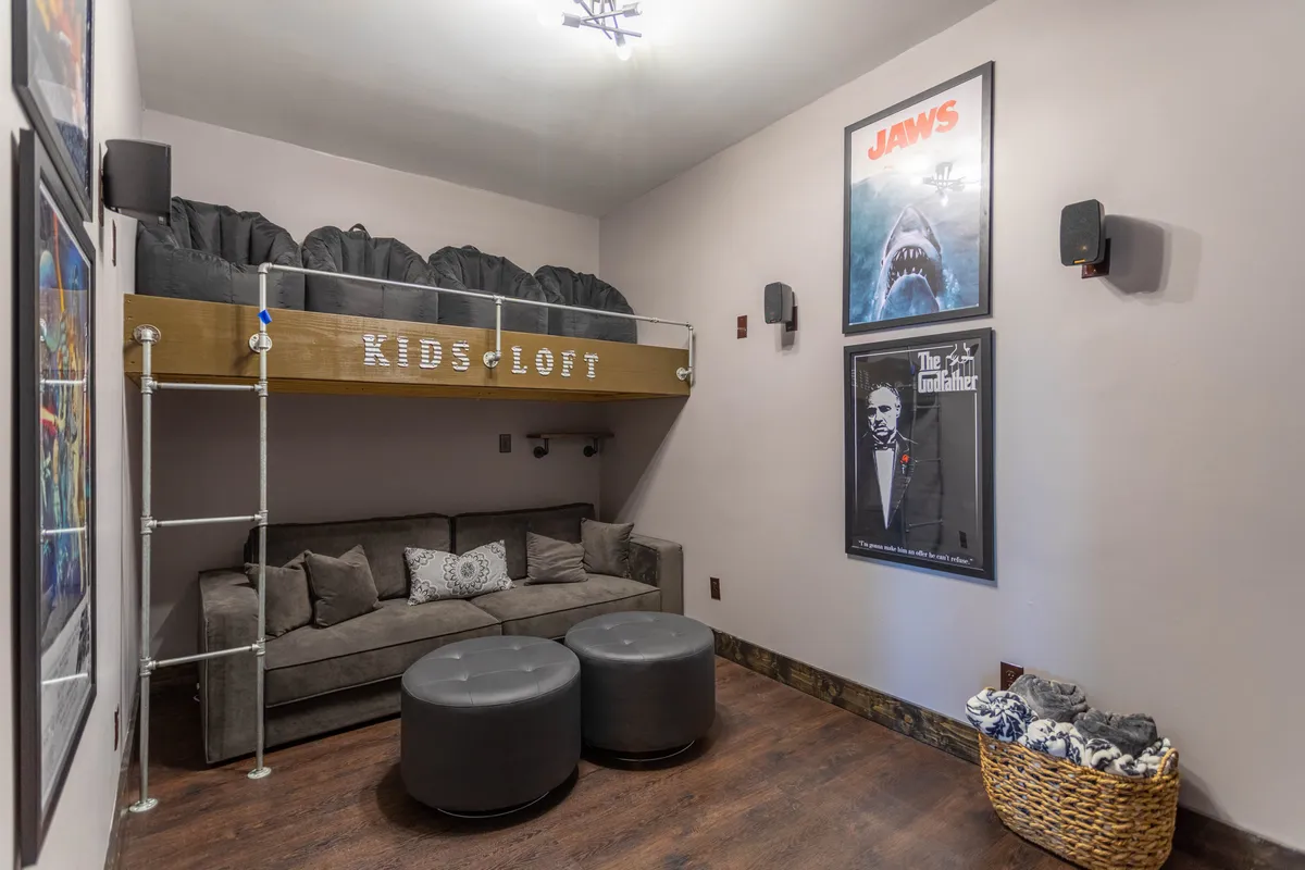A playful, family-oriented theater loft with a lofted bed, plush sofa, movie posters, ottomans, and a basket of blankets, offering a whimsical and comfortable viewing space especially suited for kids and creative play.