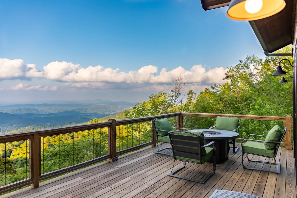 An expansive wooden deck with comfortable outdoor seating, set against a breathtaking backdrop of rolling mountains. The area features a cozy fire table, perfect for group gatherings, and ample space for relaxing or sharing stories under the sky.