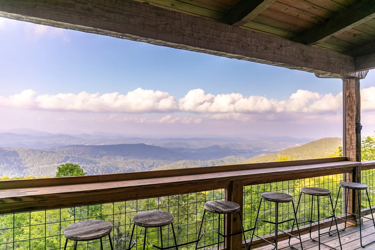 A wooden deck featuring a countertop railing with barstools, overlooking a majestic mountain landscape. The setting is ideal for enjoying morning coffee or an evening drink while taking in sunrise or sunset views.