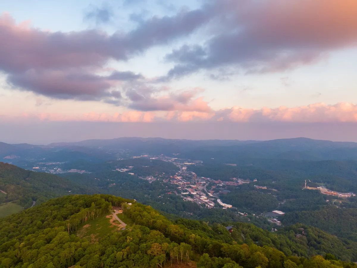 A panoramic view reveals a vast mountainous landscape, a quaint town nestled in the valley, and a sky streaked with the colorful hues of sunset. The scene evokes a sense of adventure, exploration, and awe at the natural beauty that defines the High Country region.